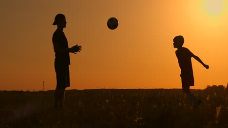 Vater-Und-Sohn-Spielen-Fußball-Im-Park-Bei-Sonnenuntergang,-Silhouetten-Vor-Dem-Hintergrund-Einer-Zeitlupenaufnahme-Einer-Hellen-Sonne.