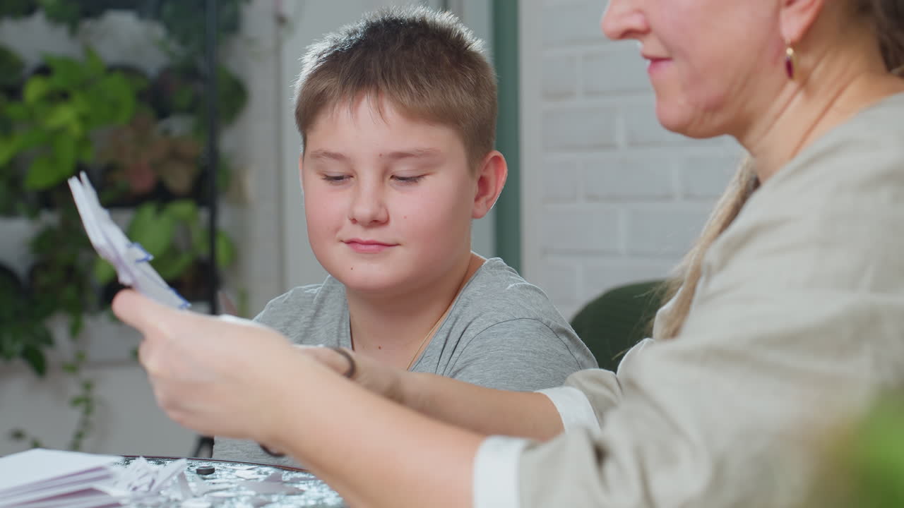 partial view of woman carving paper with scissors while boy in grey top watches closely, focused on delicate paper craft activity at home, creative bonding moment captured in bright indoor setting