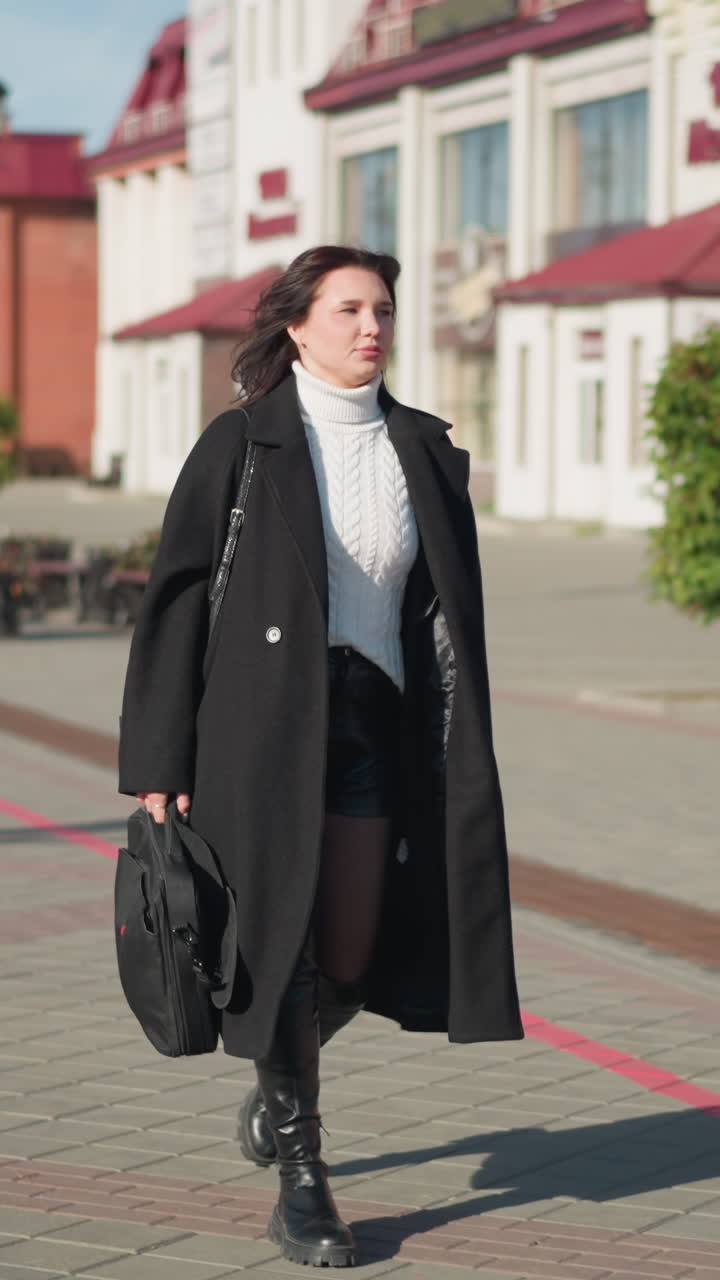 Business lady going to her office in black coat, and black boots on paved urban street during sunny day, holding black handbag with someone blur in background, surrounded by modern buildings