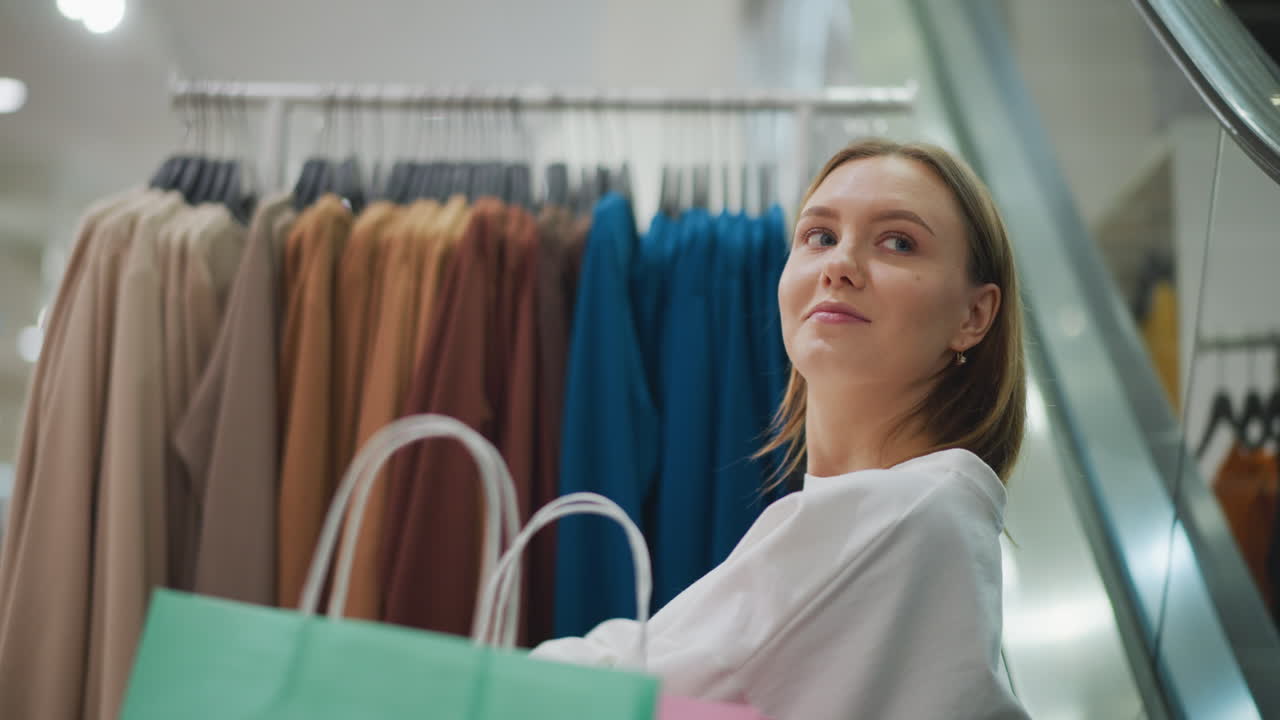 밝고 잘 조명 된 가게에서 따뜻한 미소로 옷장을 바라보는 여인이 배경에서 무언가를 관찰하고, 그녀의 근처에 쇼핑 가방을 바라보고 있습니다.