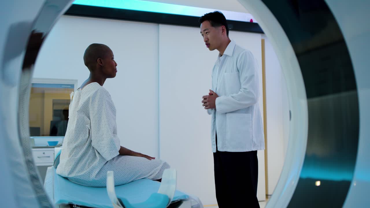 Door opening African woman gowned sitting in gantry, listening as Asian doctor entering MRI prep