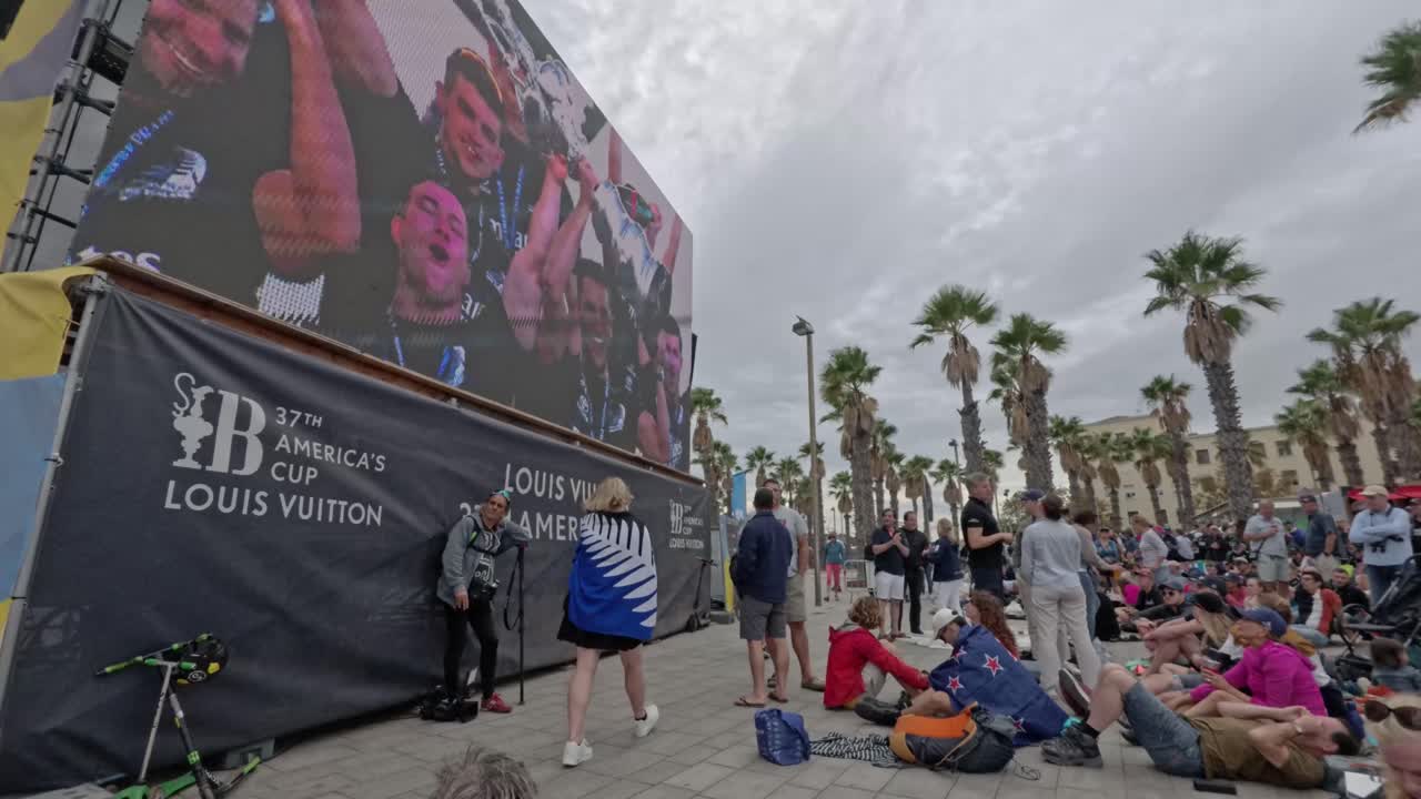 Crowd outside watching the 37th America´s cup on a giant screen. Celebrations. Palm trees in the background
