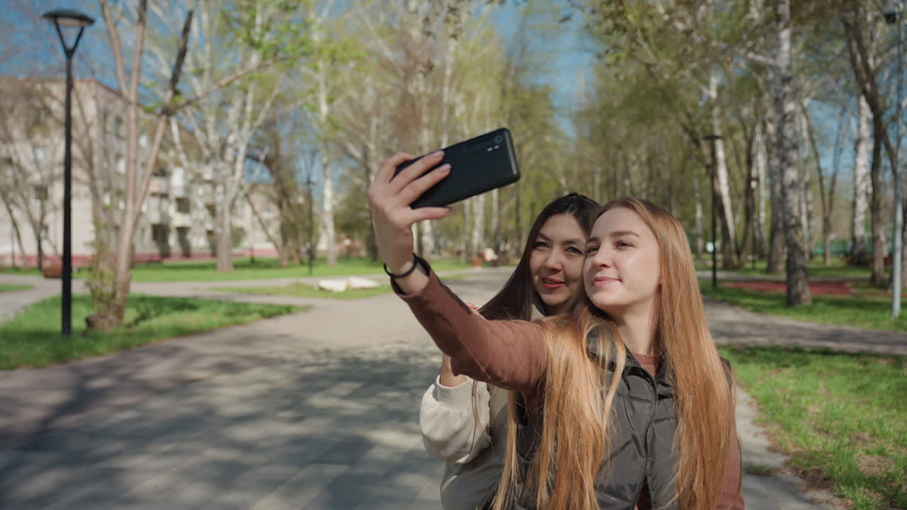selfi en un parque, retrato, caucásico, asiático, sonriendo, primer plano, luz cálida y fondo frondoso, dos amigos encuadrando sus caras con un teléfono, chaquetas informales y expresiones alegres, ambiente de creación de contenido espontáneo