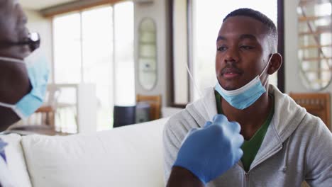 African-american-senior-male-doctor-giving-covid-test-to-male-patient-in-home,-wearing-face-masks