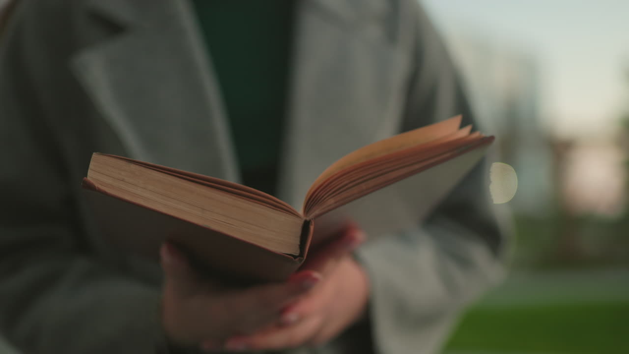 Close up of woman in gray coat holding open book with hands visible, reading attentively outdoors with blurred greenery and warm daylight creating calm intellectual atmosphere