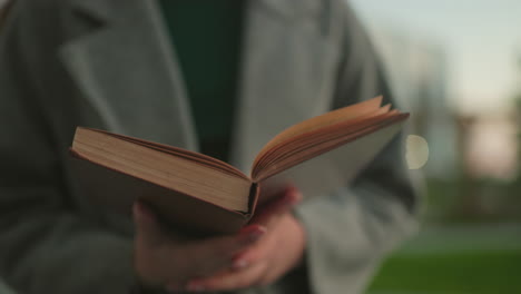 Close up of woman in gray coat holding open book with hands visible, reading attentively outdoors with blurred greenery and warm daylight creating calm intellectual atmosphere