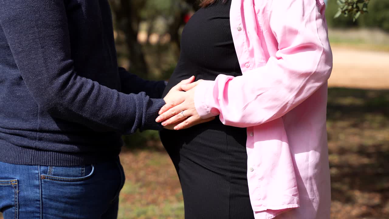 Couple stands close, hands on pregnant belly, feeling baby together outdoors