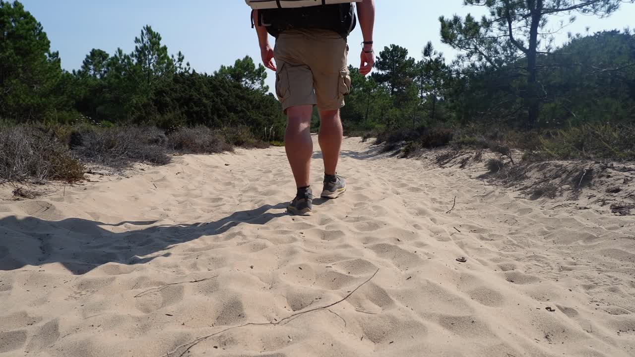 Caucasian male backpacker walks in deep sand past low angle camera