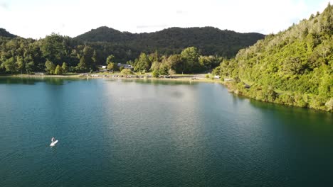 Lakeside-holiday-campground,-blue-lake,-palm-forest-road-and-tourist-on-paddle-bord-4k-drone-shot-in-New-Zealand