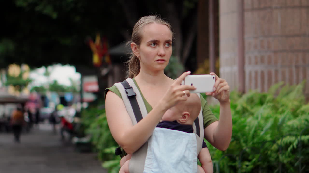 A young mother with her baby in a kangaroo backpack documents their journey through photos on a mobile phone. As she walks, she glances at the phone screen
