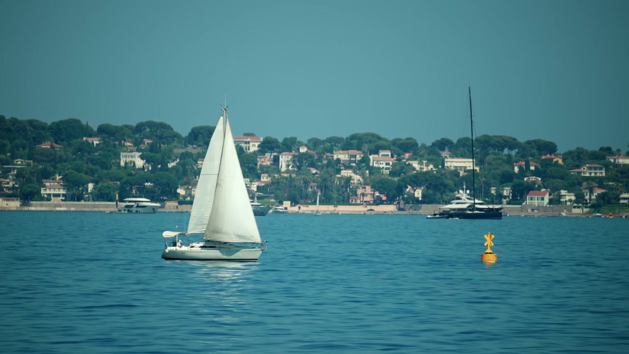Güneşli Fransız Rivierası kıyısında, Golfe Juan, Fransa'da, lüks yatlar ve kıyı villaları eşliğinde sakin mavi sularda seyreden beyaz bir yelkenli.