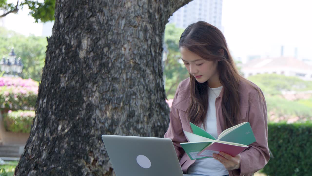 En ung kvinne leser en bok mens hun sitter med en laptop i en solrik park.