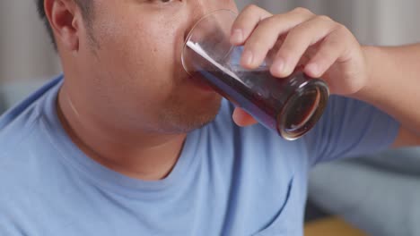 close up of a fat asian man drinking a glass of coca cola while eating fast food having fried chicken on sofa in the living room at home