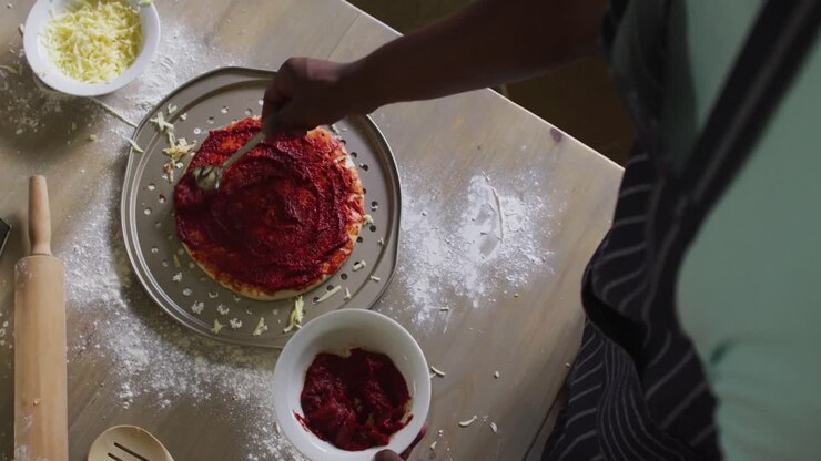 African american girl making pizza spreading tomato on base in kitchen