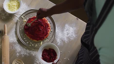 african american girl making pizza spreading tomato on base in kitchen