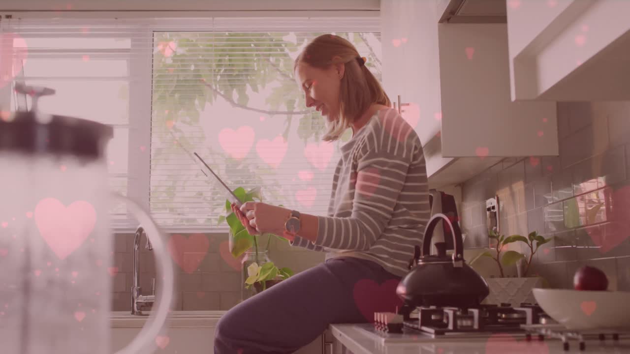 animación de emojis de corazón sobre una mujer caucásica feliz usando una tableta en la cocina