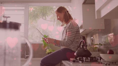 animation of heart emojis over happy caucasian woman using tablet in kitchen