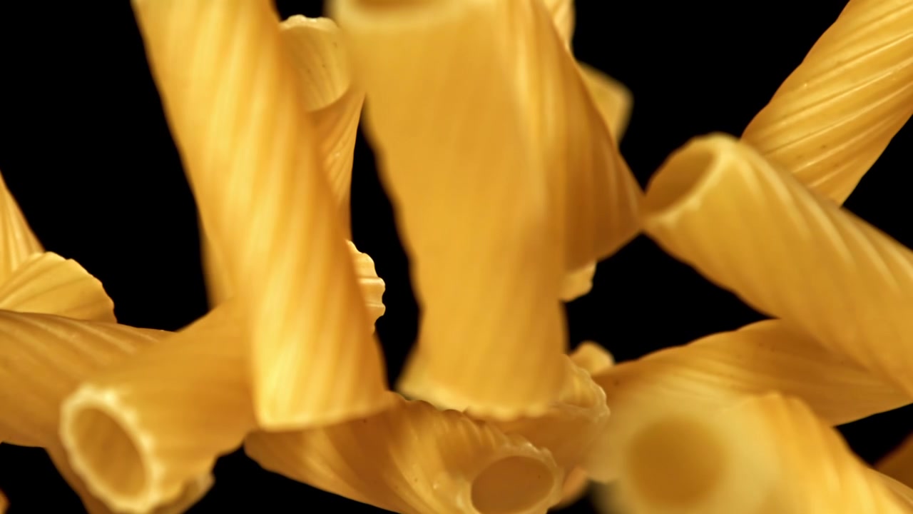 Pasta tubolare secca che cade e si solleva al rallentatore su sfondo nero.