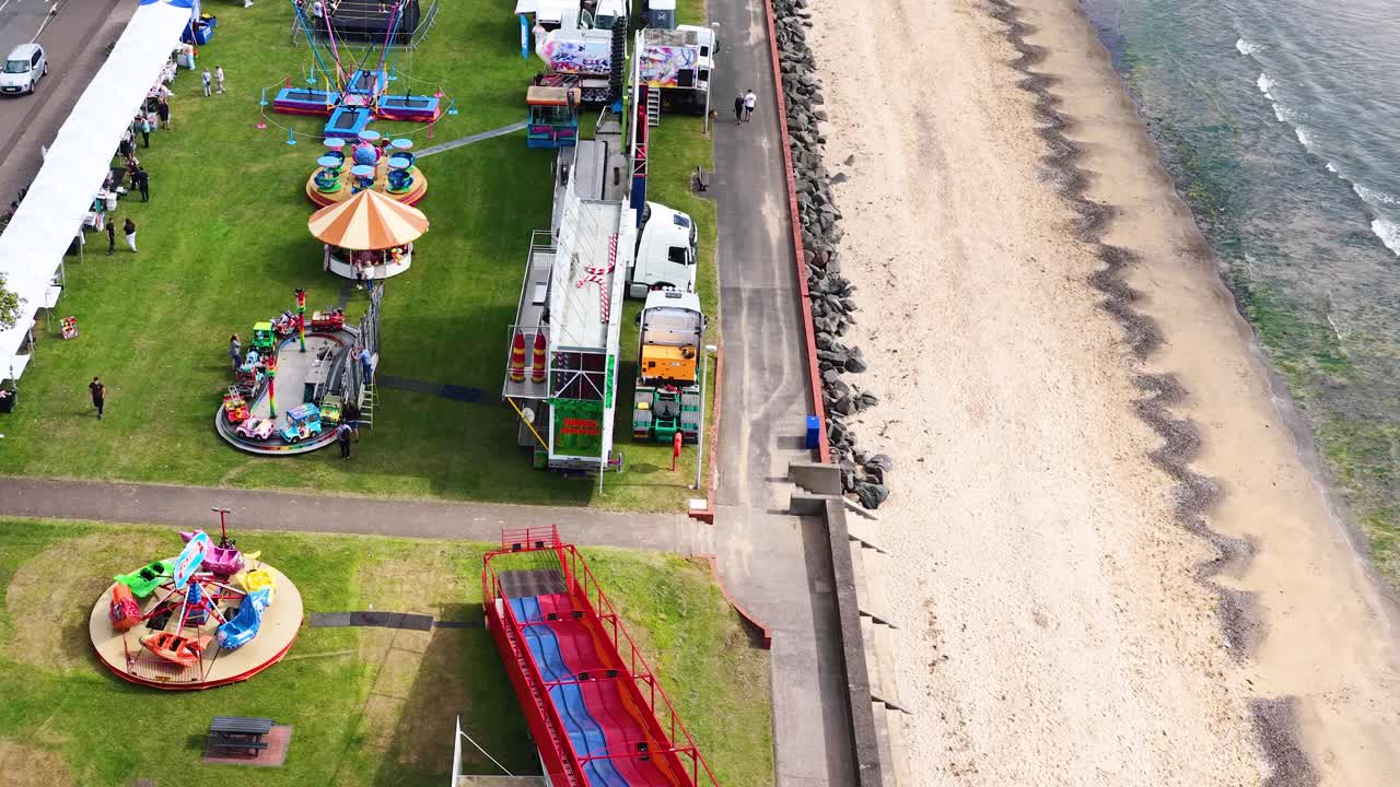 Drone ascends over seaside fairground with rides, tents, and adjacent sandy beach in daylight