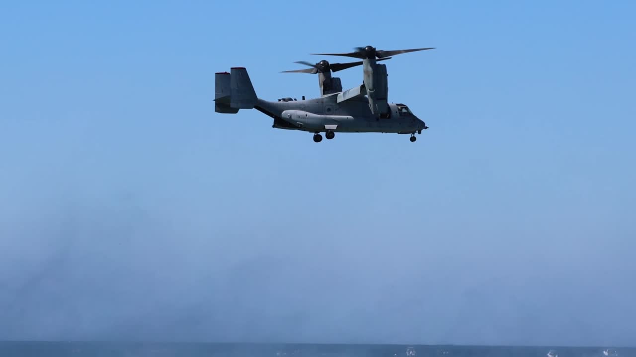 A tiltrotor aircraft hovers steadily above the ocean, with clear skies and distant waves visible.