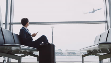 businessman sits in airport departure lounge using mobile phone with plane taking off in background