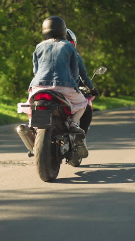 dos mujeres montando una bicicleta eléctrica, el pasajero sosteniendo firmemente al jinete, mientras viajan por una carretera sombreada bordeada de árboles, el viento aleta el vestido del pasajero, con un coche lejano visible delante