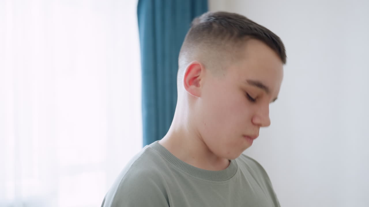 Teen boy practices neck flexibility routine in bright home setting near window with soft daylight, standing still in relaxed posture, he tilts head gently to stretch neck muscles for mobility