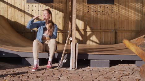 video of happy caucasian female skateboarder resting and using smartphone