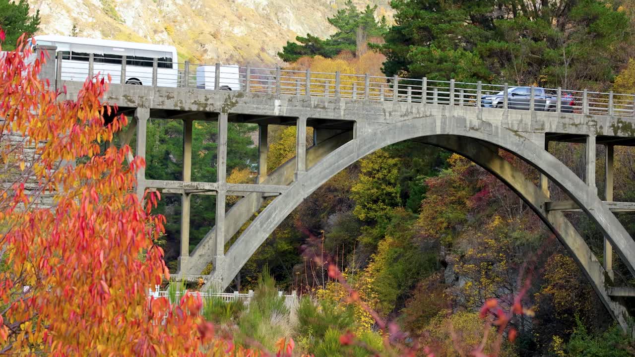 Vehicles traverse the scenic Edith Cavell Bridge in Queenstown, surrounded by vibrant autumn foliage and natural landscapes