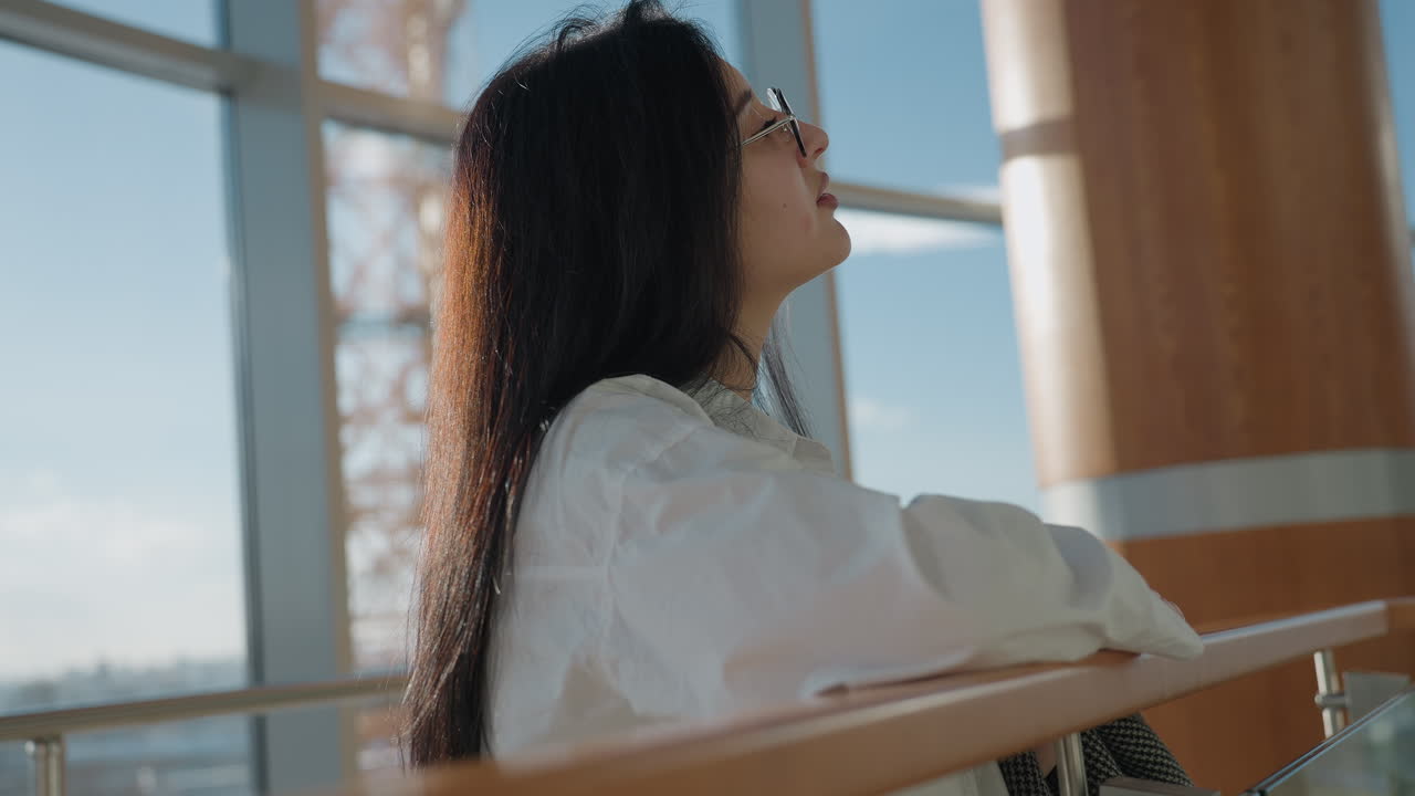 Elegant woman in white shirt and glasses rests on wooden rail bathed in natural light, gazing upward with calm thoughtful expression, long dark hair glowing in sunlight inside modern interior setting