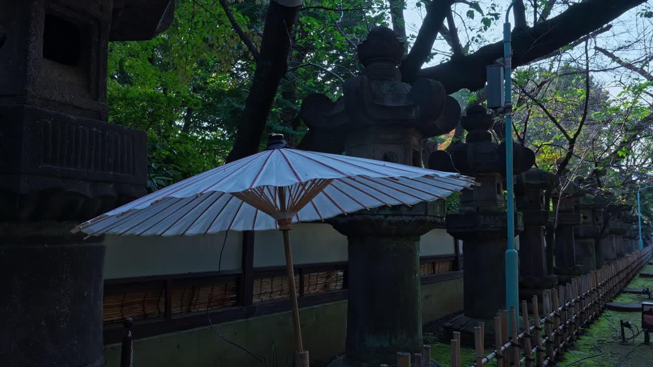 A traditional Japanese paper parasol next to ancient stone lanterns, surrounded by green trees, creating a timeless and tranquil scene.