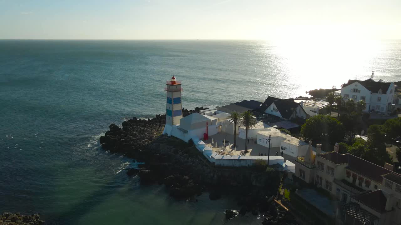 Aerial drone video flying around gorgeous and historic lighthouse called Forte de Santa Marta during a sunny day while ocean turquoise sea water waves hit the rocky shore cliffs below. Sun rays, flare
