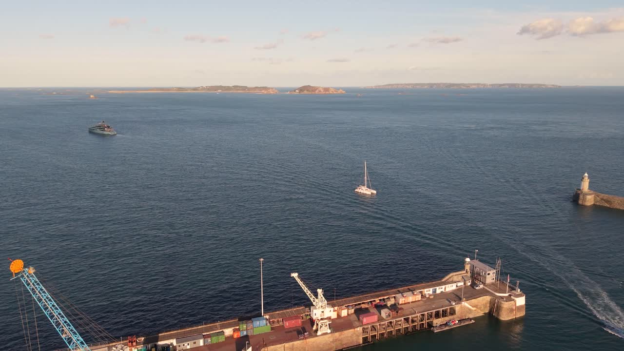 Docks and entrance to St Peter Port Harbour Guernsey with boats entering views to Herm Jethou and Sark and sun lighting up the docks and harbour wall in golden hour sunshine on calm day