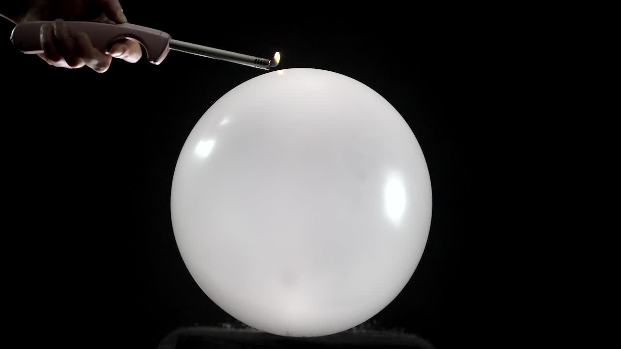 A round balloon pops in slow motion as a hand holding a lighter gets closer. The hand is visible in front of a black background, adding a dramatic effect to the tense moment before the burst.