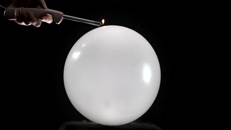 a round balloon pops in slow motion as a hand holding a lighter gets closer. the hand is visible in front of a black background, adding a dramatic effect to the tense moment before the burst.