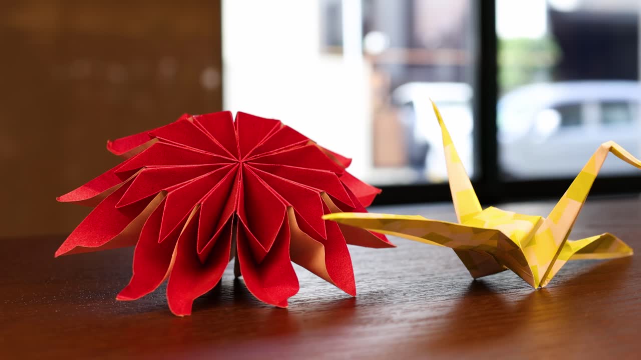 Close-up of a red origami flower and yellow crane on a wooden surface.