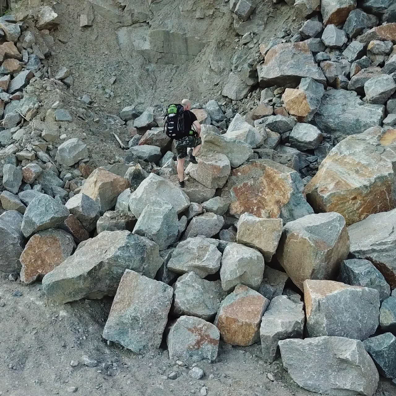 Active tourist climbing on a big pile of stones in the mountainous area. Scenic view of rocky cliff and a young man hiker walking on that place. Camera moves up.
