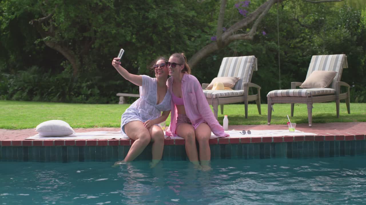 Two female friends in swimwear splashing water, sharing smartphone and taking selfies poolside