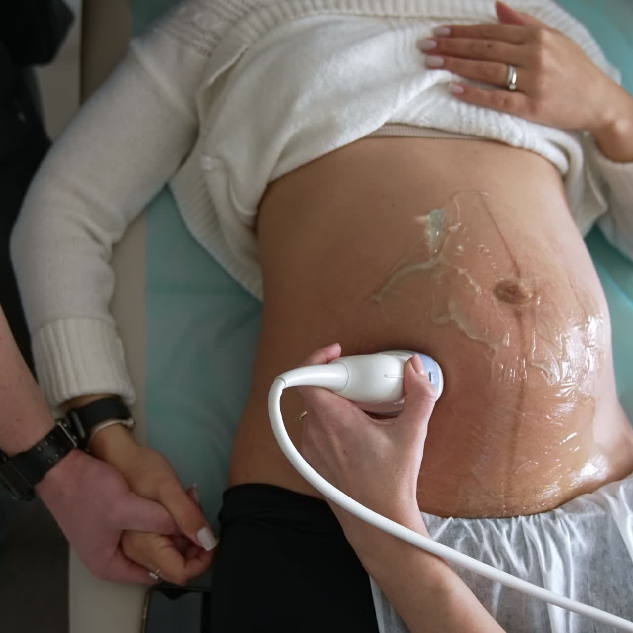 Pregnant lady lies on the doctor's couch and her husband sits beside. Obstetrician's hand moves the sensor by the big belly of expecting woman