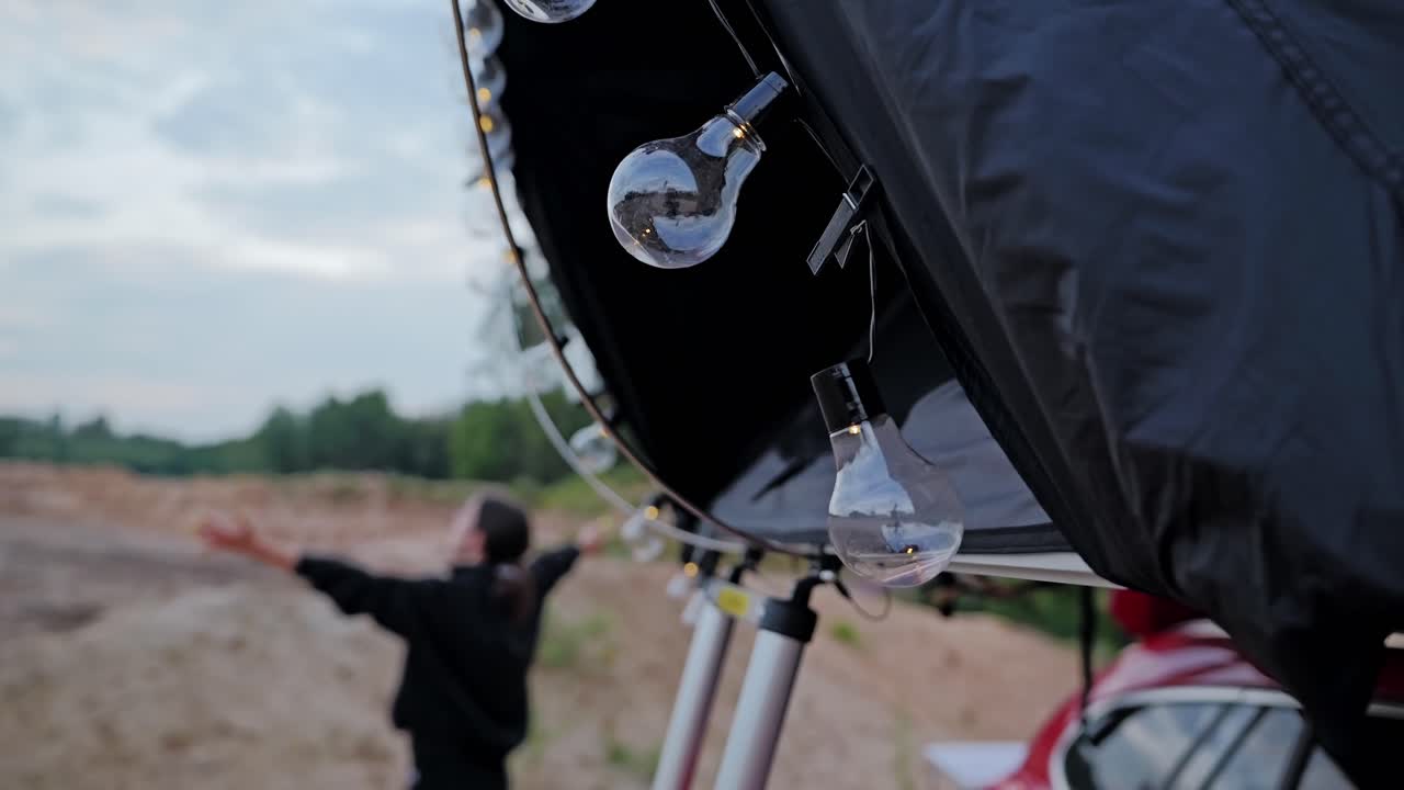 Slow motion of woman raising arms toward sky beside tent lit by string lights