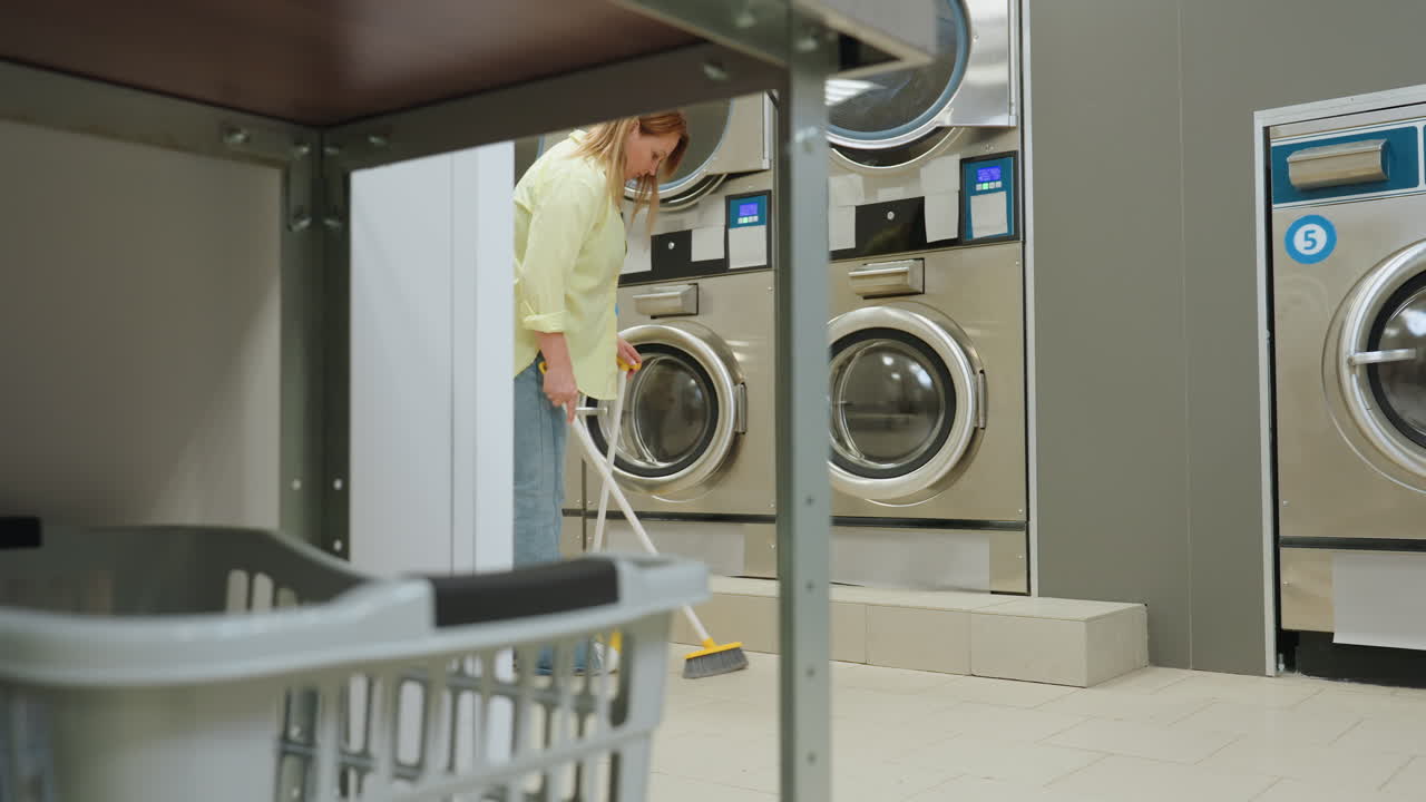 View through shelf shows laundry cleaner sweeping floor beside industrial washers with broom and dustpan, keeping tidy service facility, stainless machines, tiled room, janitorial routine