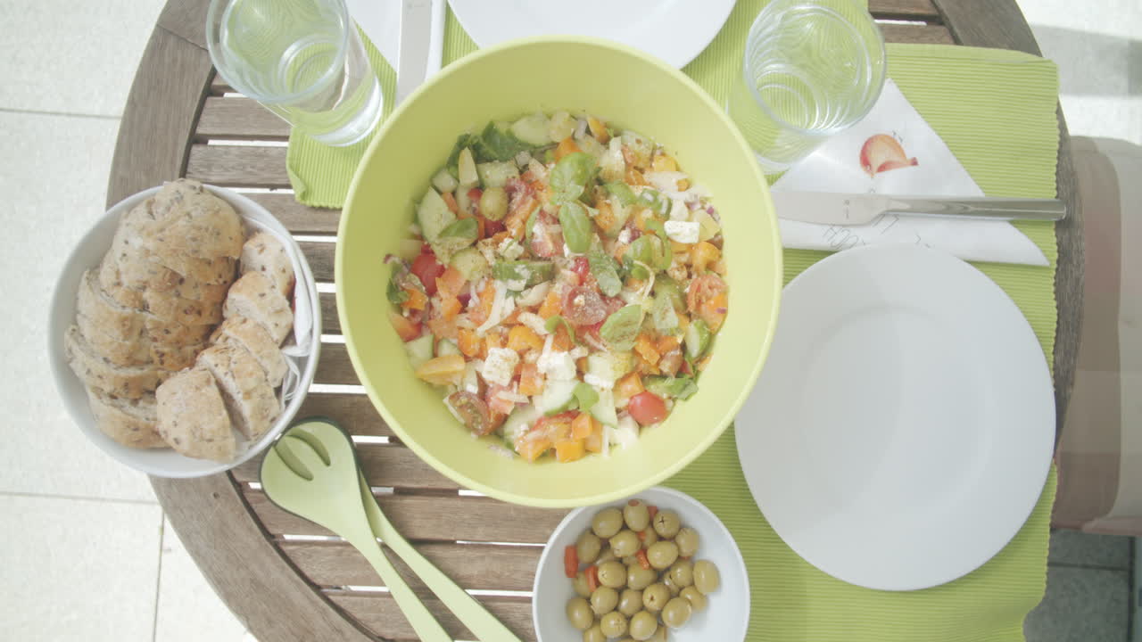 Top shot of a laid table on a sunny spring day. Caucasian hands putting food onto the table. Bowl filled with greek salad, another bowl with bread and a small bowl with olives