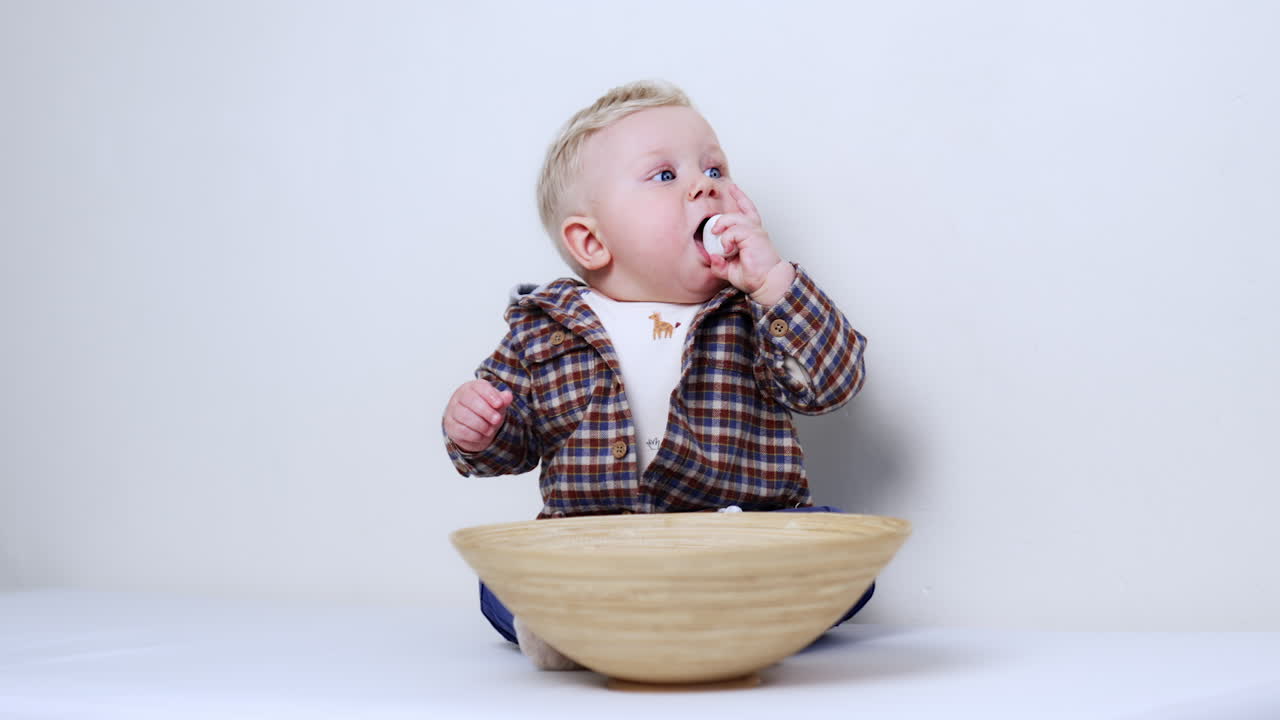 Little baby boy in checkered shirt sits on the floor with a bowl in front of him. Kid takes a white pebble from the dish and shoves it to his mouth.