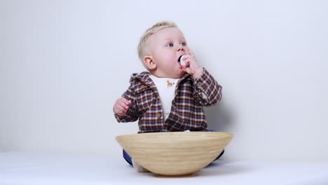 little baby boy in checkered shirt sits on the floor with a bowl in front of him. kid takes a white pebble from the dish and shoves it to his mouth.