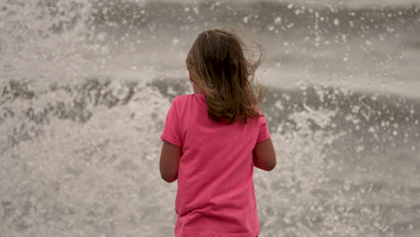 las olas salpican a una niña mientras está de pie en una playa mirando el océano