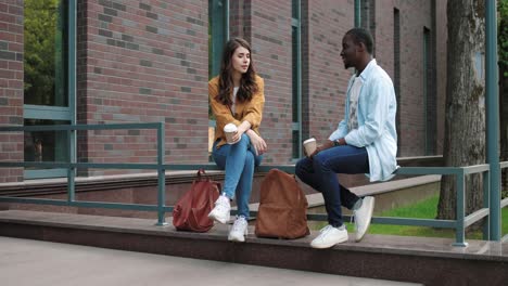 camera zooming on caucasian and african american students talking and drinking coffee sitting in the street near the college