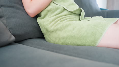 close view of person wrapped in green towel lowering body to sit on gray cushioned couch, focusing on towel and soft furniture texture in calm indoor environment with minimal background distraction