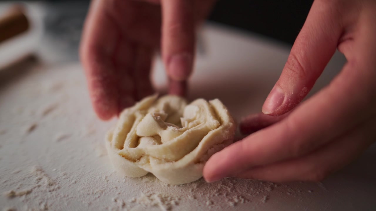 Close-up of hands shaping dough into a rose. Fingers gently mold the soft, pale dough. Dusting flour adds texture to the workspace. Calm, focused action suggests baking or culinary art