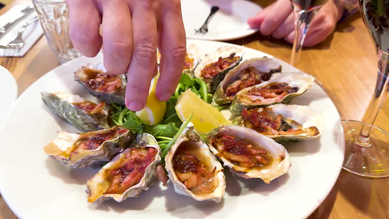 A hand squeezes lemon over a plate of oysters, enhancing their flavor. The setting is a well-lit dining table