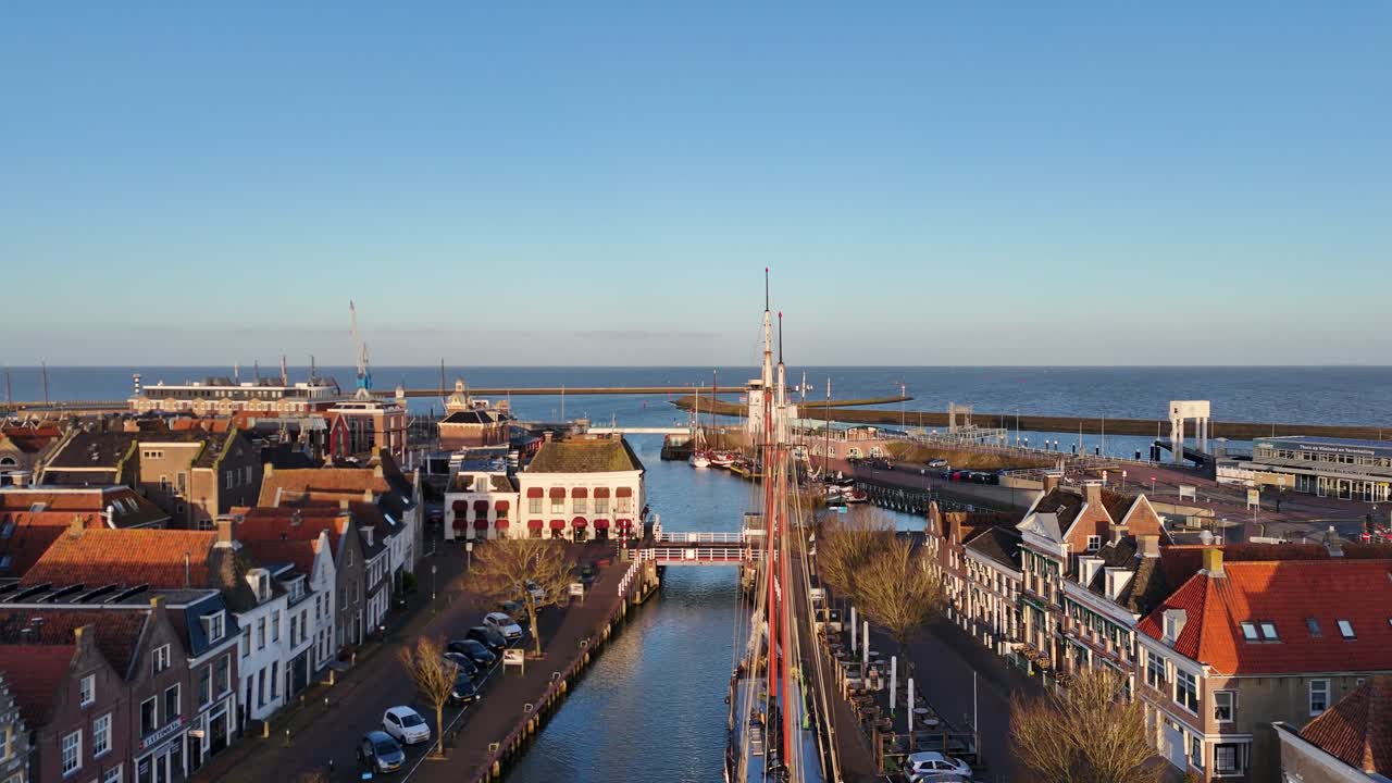 Eine Drohne sinkt auf ein großes Segelschiff zu, das in einem engen Kanal am Hafen festgemacht ist.